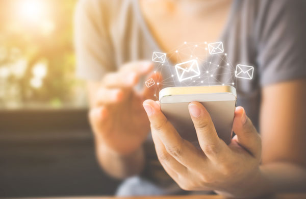 Osoba trzyma smartfona, wokół którego unoszą się ikony kopert, symbolizujące wysyłanie wiadomości e-mail.
