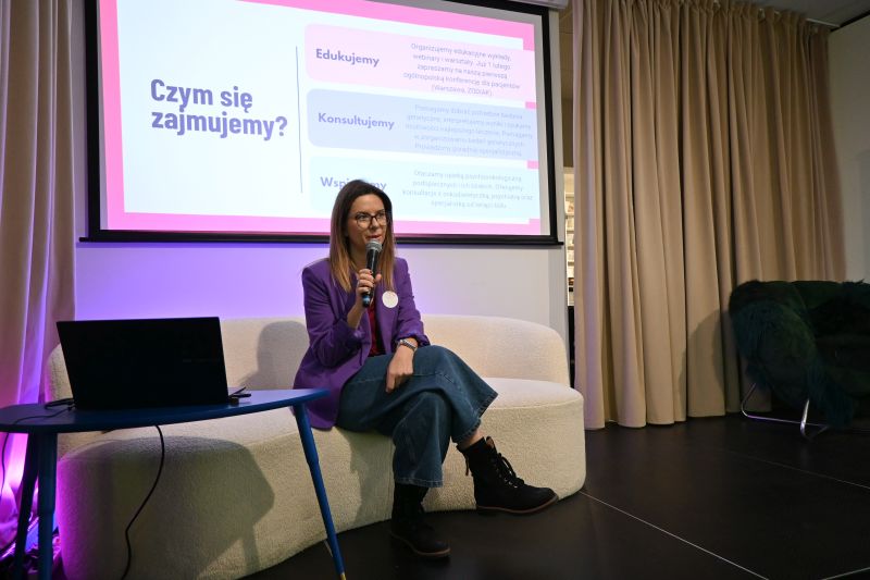 Kobieta w fioletowej marynarce siedzi z mikrofonem przed ekranem z prezentacją Czym się zajmujemy?.