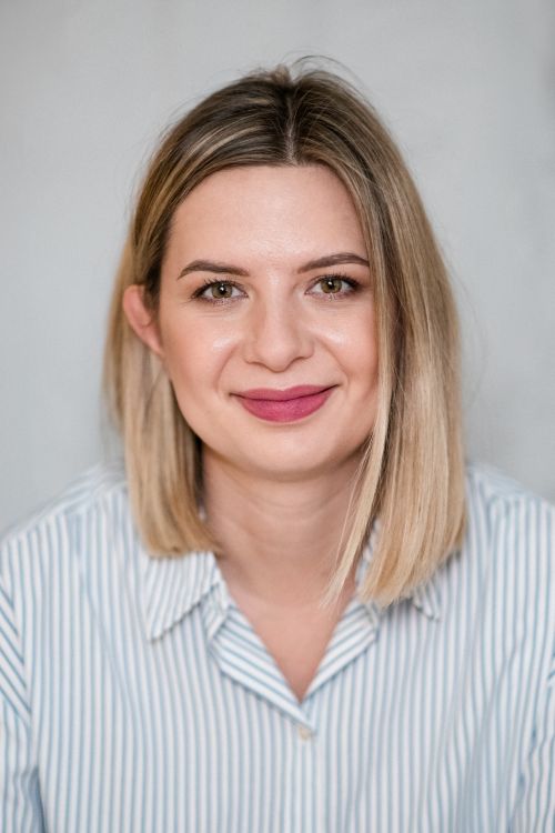 Kobieta z krótkimi blond włosami uśmiecha się, ubrana w koszulę w paski, na neutralnym tle.