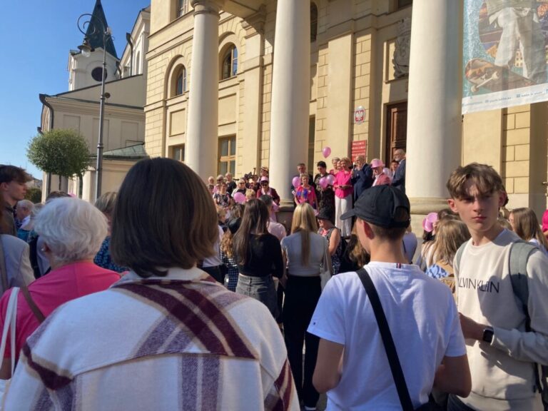 Grupa ludzi zgromadzona przed zabytkowym budynkiem, część ubrana na różowo, prowadzi wydarzenie lub protest.