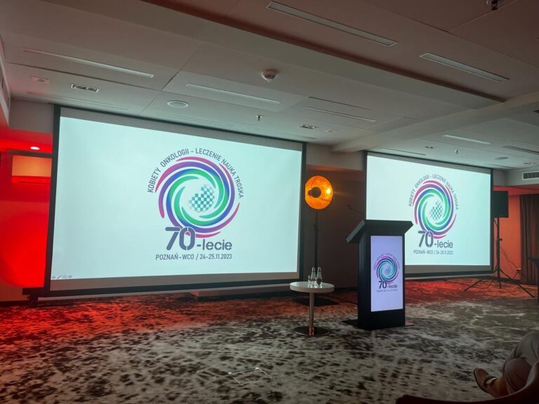 Sala konferencyjna z ekranem projekcyjnym, logo 70-lecie Kobiety Onkologii oraz podium.