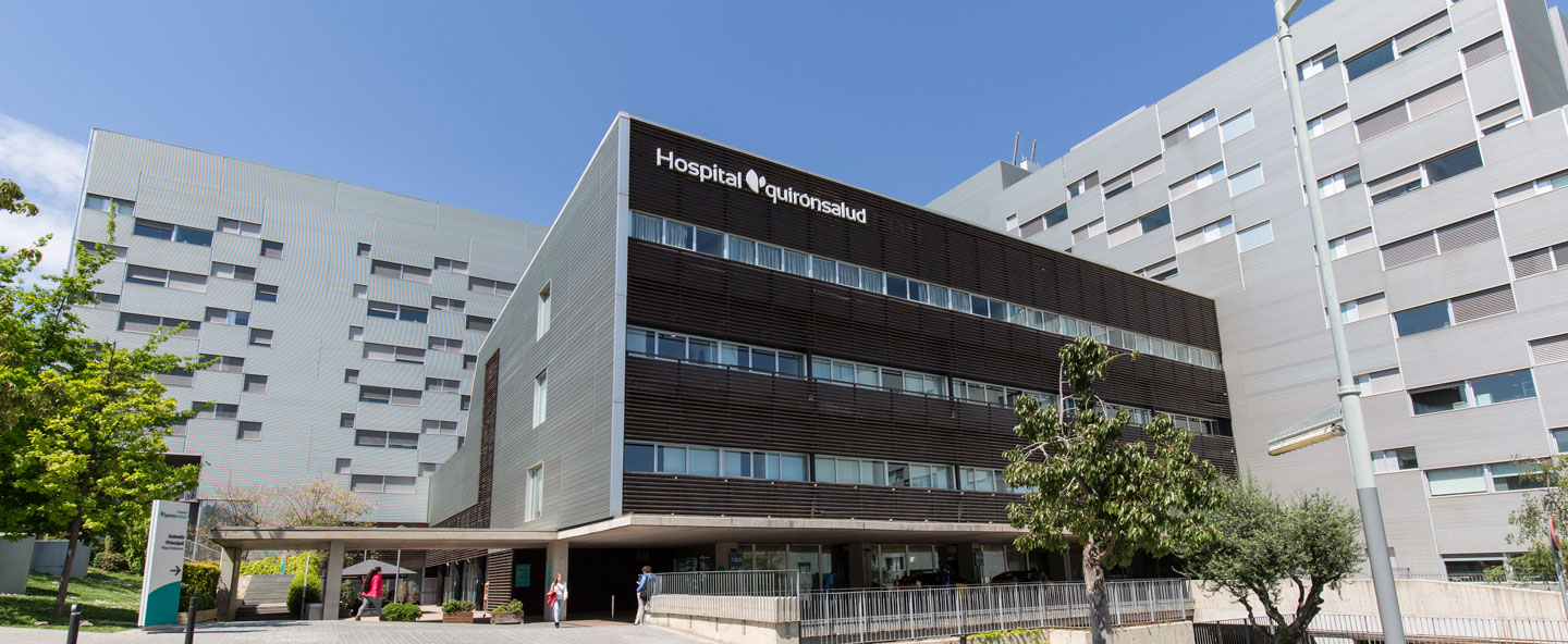 Nowoczesny budynek szpitalny, Hospital Quirónsalud, w otoczeniu zieleni, na tle jasnego nieba.