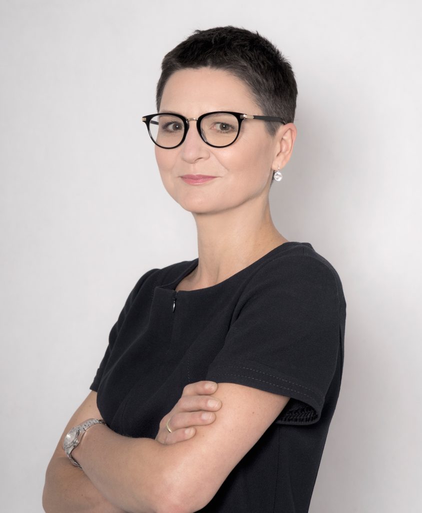 Osoba w okularach z krótkimi włosami i krzyżującą ramiona, ubrana w ciemną bluzkę, na jasnym tle.