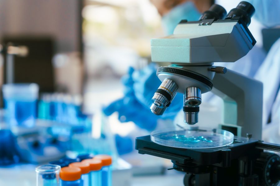 Mikroskop laboratoryjny z preparatami do analizy, w tle probówki z niebieskim płynem.